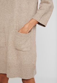 Abito lavorato a maglia beige con maniche lunghe, due tasche frontali, polsini a coste e orlo arrotondato, che mette in mostra una morbida texture e un design casual.