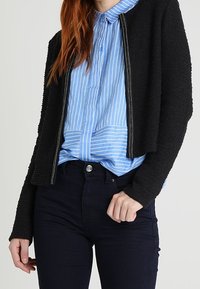 Černá texturovaná bunda s krátkým střihem a stříbrným zipem, přes světle modrou pruhovanou košili s knoflíky. K outfitu se hodí tmavé džíny.