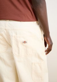 Pantalons cargo beiges avec deux poches arrière, arborant un petit empiècement en logo. Matière en coton douce avec une coupe décontractée et des détails de couture visibles.