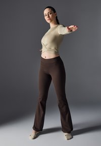 Beige langermet topp med samlet midje, sammen med brune tettsittende flared leggings. Modellen poserer med en arm strukket ut; enkel bakgrunn.