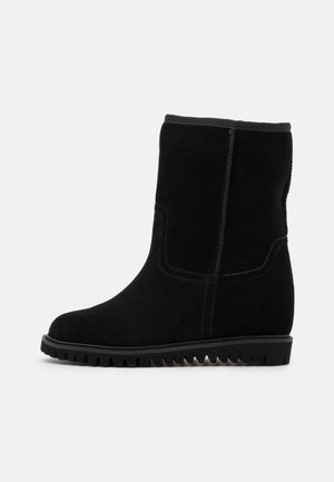 Botte noire en daim mi-mollet avec bout arrondi, couture visible et semelle épaisse en caoutchouc avec rainures profondes pour l’adhérence.