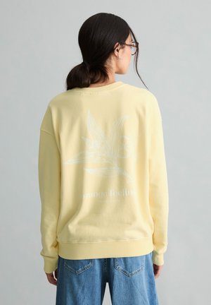 Frau mit dunklen Haaren in einem niedrigen Pferdeschwanz, die eine Brille, ein blassgelbes Sweatshirt mit Blumenmuster und Text auf dem Rücken sowie blaue Jeans trägt.