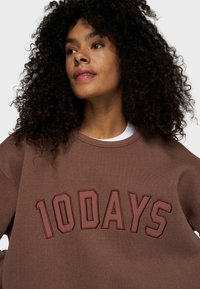 Brauner Pullover mit gesticktem "10DAYS" in dunklerem Braun. Er hat einen runden Hals und eine lockere Passform, mit strukturiertem Stoff und sichtbaren Nähten.