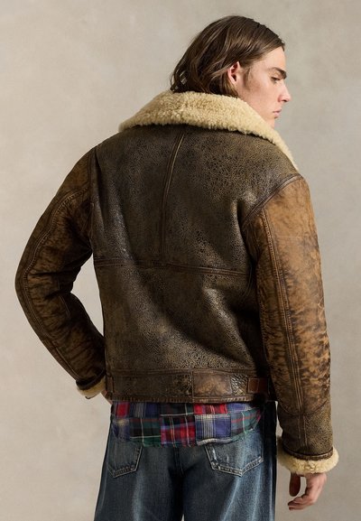 Giovane con capelli alla lunghezza delle spalle che indossa una giacca di pelle marrone con interno in shearling, camicia a quadri e jeans blu, visto di profilo con la schiena per lo più visibile.