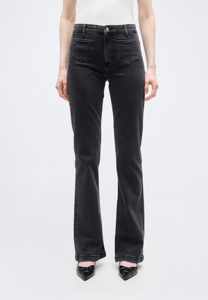 Frau trägt hoch taillierte schwarze Jeans mit geradem Bein und schwarze spitze Schuhe, steht vor einem einfarbigen hellen Hintergrund.
