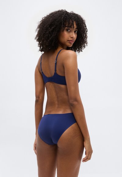 Γυναίκα με κατσαρά μαλλιά που φοράει μπλε σουτιέν και ασορτί εσώρουχα, κοιτάζοντας πάνω από τον ώμο της με φόντο ένα απλό φωτεινό σκηνικό.