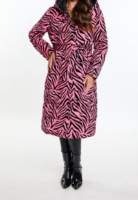 Różowo-czarna kurtka puchowa w wzór zebry z kapturem, wiązana w talii, z puszystą pikowaną fakturą. Noszona na czarnych legginsach i błyszczących botkach za kostkę.