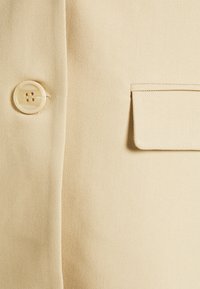 Tissu beige avec un bouton à quatre trous à gauche et une poche à rabat rectangulaire sur le côté droit.