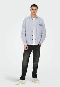 Stripet skjorte med knapper i blå og hvid, med en brystlomme og blå broderi. Båret over en hvid skjorte, parret med sorte jeans.