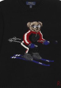 Svart stickad tröja med en björn som åker skidor, bär glasögon och en röd och svart skiddräkt. Designen inkluderar skidstavar och blå skidor.