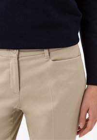 Beige op maat gemaakte broeken met een gladde textuur, ritssluiting en knoopsluiting. Zijzakdetails zichtbaar, gedragen met een marineblauwe trui.