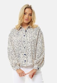 Button-up blouse in cream with dark blue heart patterns. Features a collared neck, puffed sleeves, and button front closure.