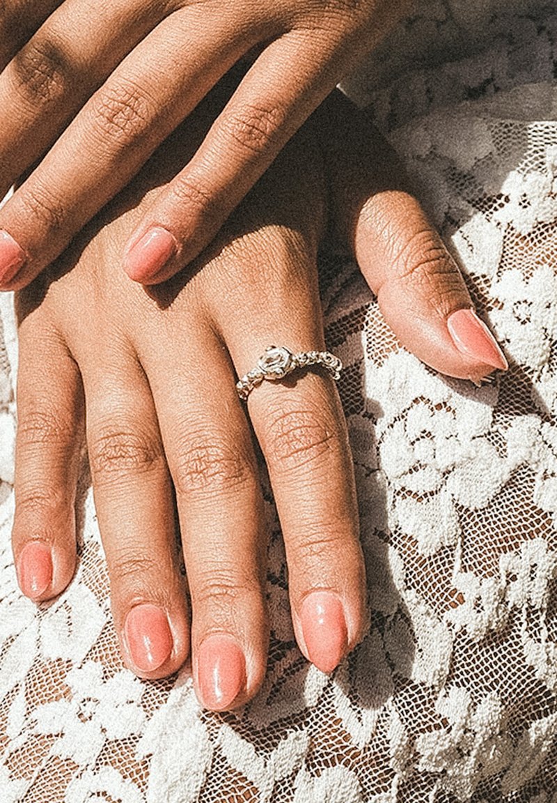 Hænder med manicurerede negle hvilende på blonder, en hånd bærer en flettet sølvring med en klar sten på ringfingeren.