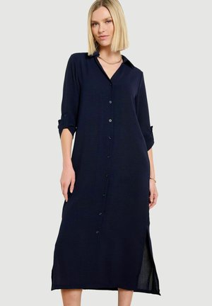 Femme aux cheveux blonds coupés au carré, portant une longue robe chemise bleu marine à boutons avec des manches retroussées aux trois quarts, sur un fond clair uni.