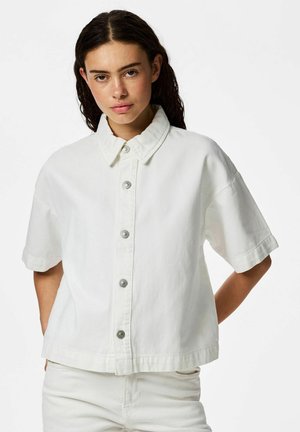 Ung kvinde med mørkt hår iført løs hvid kortærmet skjorte med knapper og hvide bukser, stående foran en ensfarvet hvid baggrund.