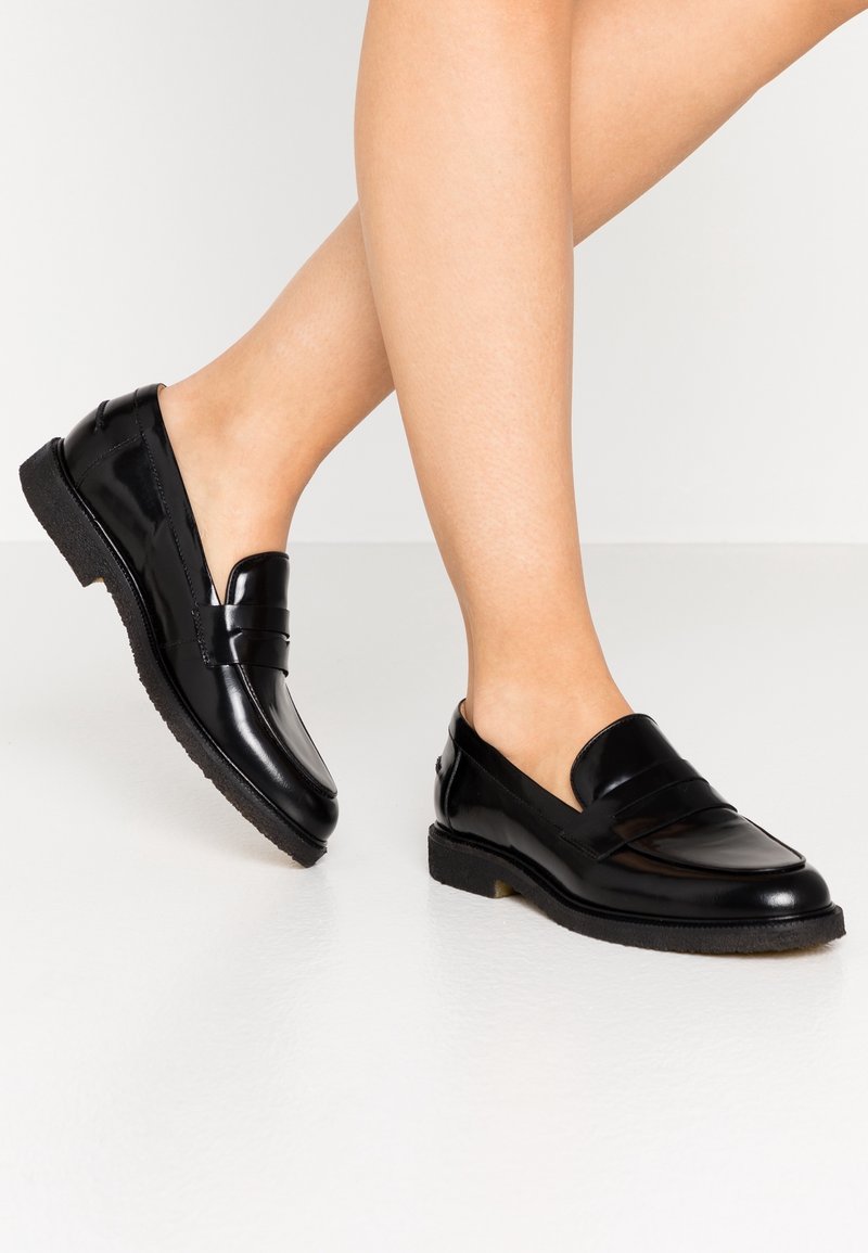 Jambes portant des mocassins en cuir noir brillant avec des semelles en caoutchouc épaisses, debout sur un sol clair contre un fond uni.