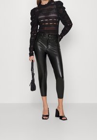 Černý krajkový top s nabíranými rukávy, kombinovaný s vysokými lesklými tmavě zelenými legínami. Modelka drží malou černou kabelku a má na sobě černé sandály na podpatku.