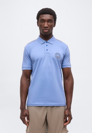 Uomo che indossa una polo azzurro chiaro con il logo del circolo tennis sul petto e pantaloncini beige, in piedi davanti a uno sfondo chiaro uniforme.