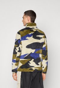 Fleecejacke mit hohem Kragen. Verfügt über ein mehrfarbiges Camouflage-Muster in Grün, Blau, Schwarz und Creme. Weicher Stoff; lockere Passform.