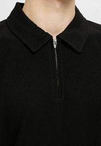 Czarny polo z prążkowanym wzorem, z zamkiem błyskawicznym z przodu i klasycznym kołnierzykiem. Tkanina ma teksturowane wykończenie z widocznymi pionowymi liniami.