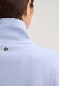 Donna con capelli scuri che indossa un top azzurro chiaro con colletto alto caratterizzato da un piccolo logo metallico rotondo sulla parte superiore della schiena.