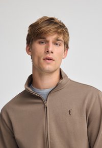 Felpa marrone con zip e colletto alto, con logo ricamato sul lato sinistro del petto, indossata sopra una t-shirt grigia chiara. Texture morbida.