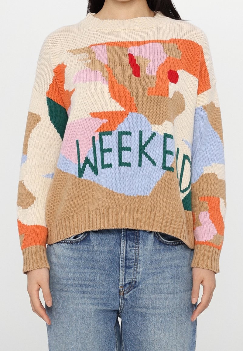Pull avec un motif abstrait multicolore et un texte vert « WEEKEND », porté avec un jean en denim bleu clair, visible du cou aux hanches.