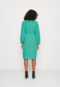 Grünes Kleid mit einem gepunkteten Muster, langen Ärmeln und taillierter Silhouette, kombiniert mit schwarzen Knöchelschuhen. Glatte Stofftextur.