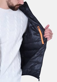 Giubbotto imbottito nero con un design a trapunta e una tasca con zip arancione, indossato sopra un maglione bianco a trecce, che mostra i dettagli interni.