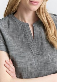 Blouse à carreaux noir et gris avec des manches courtes, un col en V et un tissu texturé, affichant un motif de grille subtil.