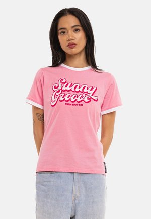Femme aux longs cheveux foncés portant un T-shirt rose avec bordure blanche et texte rouge "Sunny Groove Von Dutch" et un jean bleu clair.