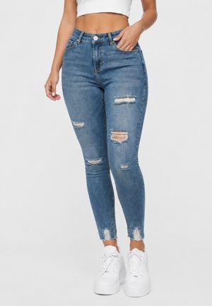 Kvinde iført højtaljede blå skinny jeans med revnede kanter og frynset kant samt hvide platformsneakers, stående foran en ensfarvet baggrund.