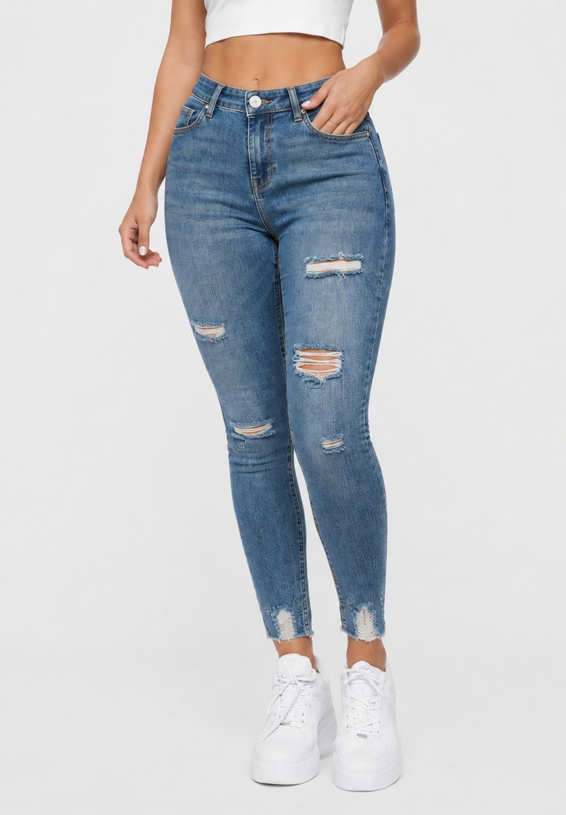Woman wearing high-waist ripped blue skinny jeans with frayed hems and white platform sneakers, standing against a plain background.