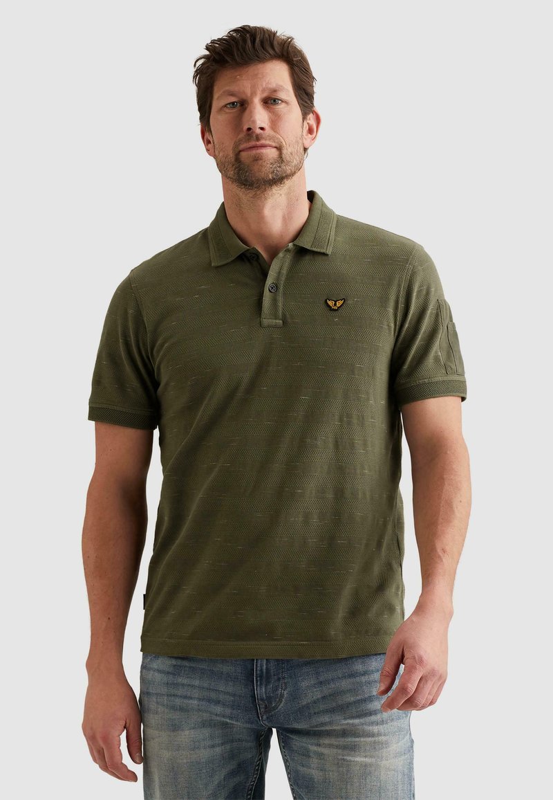 Homme portant un polo texturé vert olive avec un petit logo brodé sur la poitrine, associé à un jean bleu, debout devant un fond uni.