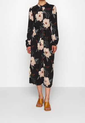 Kvinde iført en sort midikjole med stort beige blomsterprint, lange ærmer, knapper, taljebånd og brune åbne hæle-sandaler.
