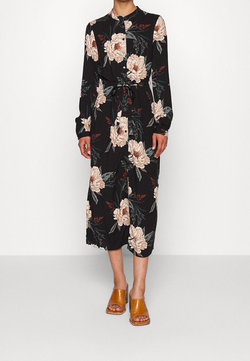Femme portant une robe midi noire avec un grand imprimé floral beige, manches longues, boutons, ceinture à la taille et sandales à talons ouvertes couleur beige.