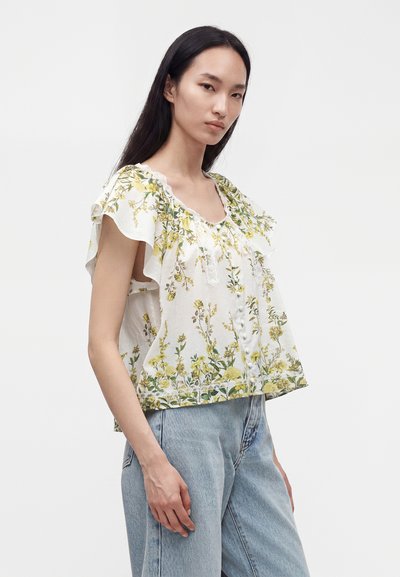 Jeune femme portant un chemisier blanc à imprimé floral jaune et manches volantées, associé à un jean bleu clair sur un fond uni.