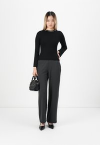 Donna con un maglione nero a maniche lunghe e pantaloni larghi scuri, che tiene in mano una piccola borsa nera, in piedi di fronte a uno sfondo bianco semplice.