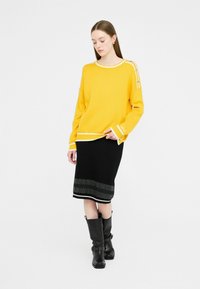 Maglione giallo lavorato a maglia con bordi bianchi, abbinato a una gonna nera con orlo decorato e stivali neri al ginocchio con fibbie.