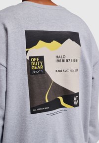 Grafika górska z żółtymi, czarnymi i beżowymi kształtami, z tekstem "OFF DUTY GEAR", "HALO 1968|972|981" oraz "ALL TERRAIN WEAR" na szarej tkaninie.