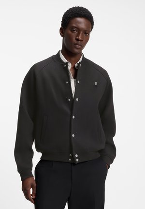 Young man wearing a black buttoned bomber jacket over a white striped shirt with hands relaxed by his sides against a white background.