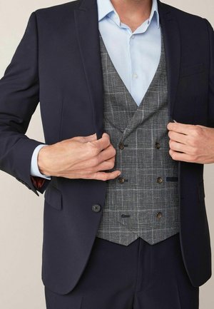 Homme ajustant une veste bleu marine par-dessus un gilet croisé à carreaux gris et une chemise habillée bleu clair.