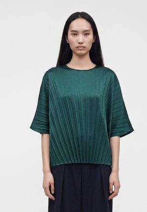 Femme aux longs cheveux foncés portant un haut plissé vert à manches courtes et un pantalon large foncé, debout devant un fond clair uni.