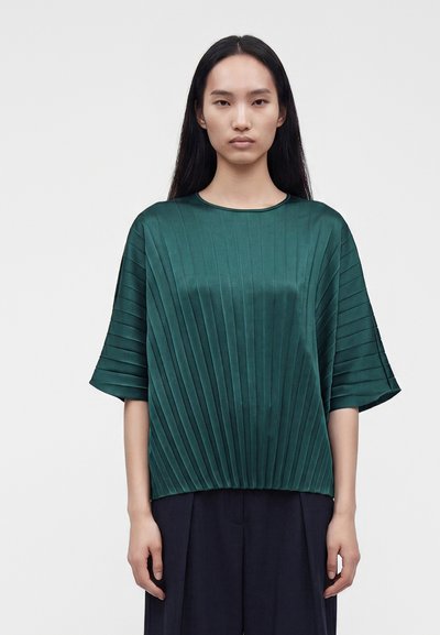 Femme aux longs cheveux foncés portant un haut plissé vert à manches courtes et un pantalon large foncé, debout devant un fond clair uni.