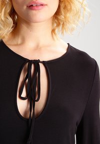 Närbild av en person som bär en svart topp med en nyckelhålsutskärning, knuten med svarta tygsnoddar, vilket visar en del av nyckelbenet och läpparna.