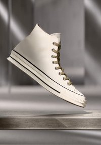 Zapatillas altas de lona beige con cordones blancos, suela de goma negra y una franja negra contrastante a lo largo del borde inferior.