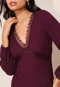 Bordó bordázott ruha mély V nyakkivágással, csipkés díszítéssel, hosszú ballon ujjakkal és testhezálló derékkal rendelkezik. A texturált anyag mélységet ad.