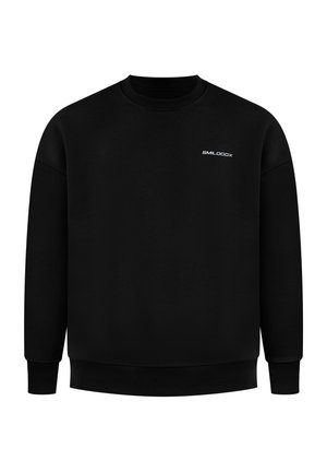 Man met een zwarte trui met een witte afbeelding op de rug en lichtgrijze joggingbroek, staand en over zijn schouder kijkend.