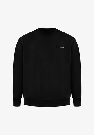 Mann trägt schwarzen Pullover mit weißem Motiv auf dem Rücken und hellgraue Jogginghose, steht und schaut über die Schulter.