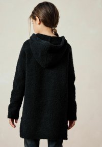 Hoodie noir en velours, au toucher doux et texturé, à manches longues et coupe décontractée, présentant une vue de dos et un ourlet légèrement descendu.